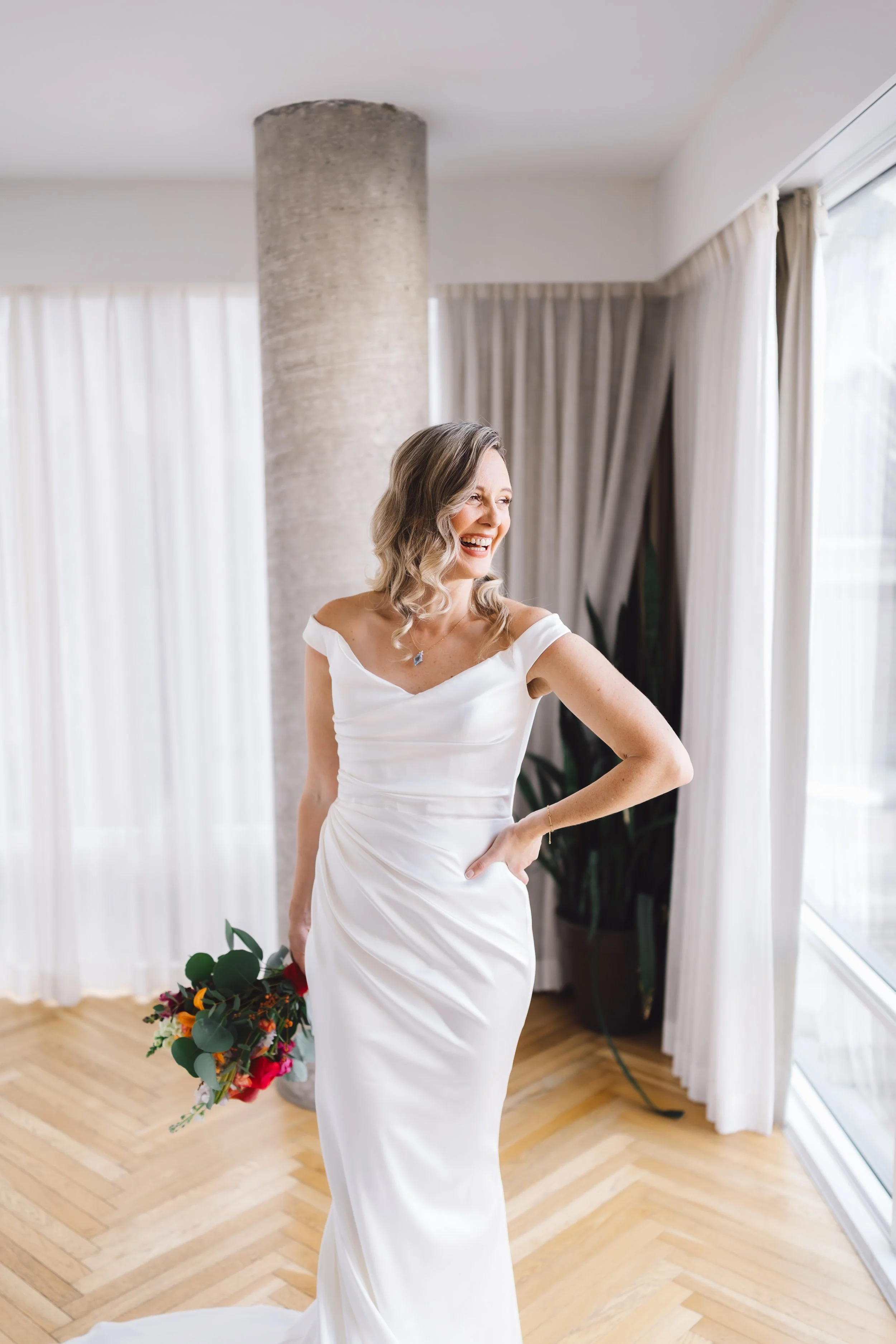A woman in a white wedding dress holding a bouquet, smiling, standing indoors near large windows with curtains.