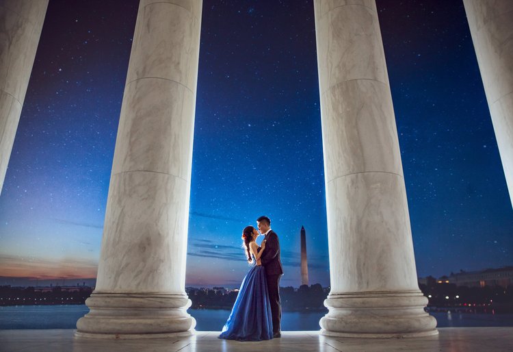 A romantic couple standing close together at night under a starry sky, framed by large classical columns with the Washington Monument in the background.