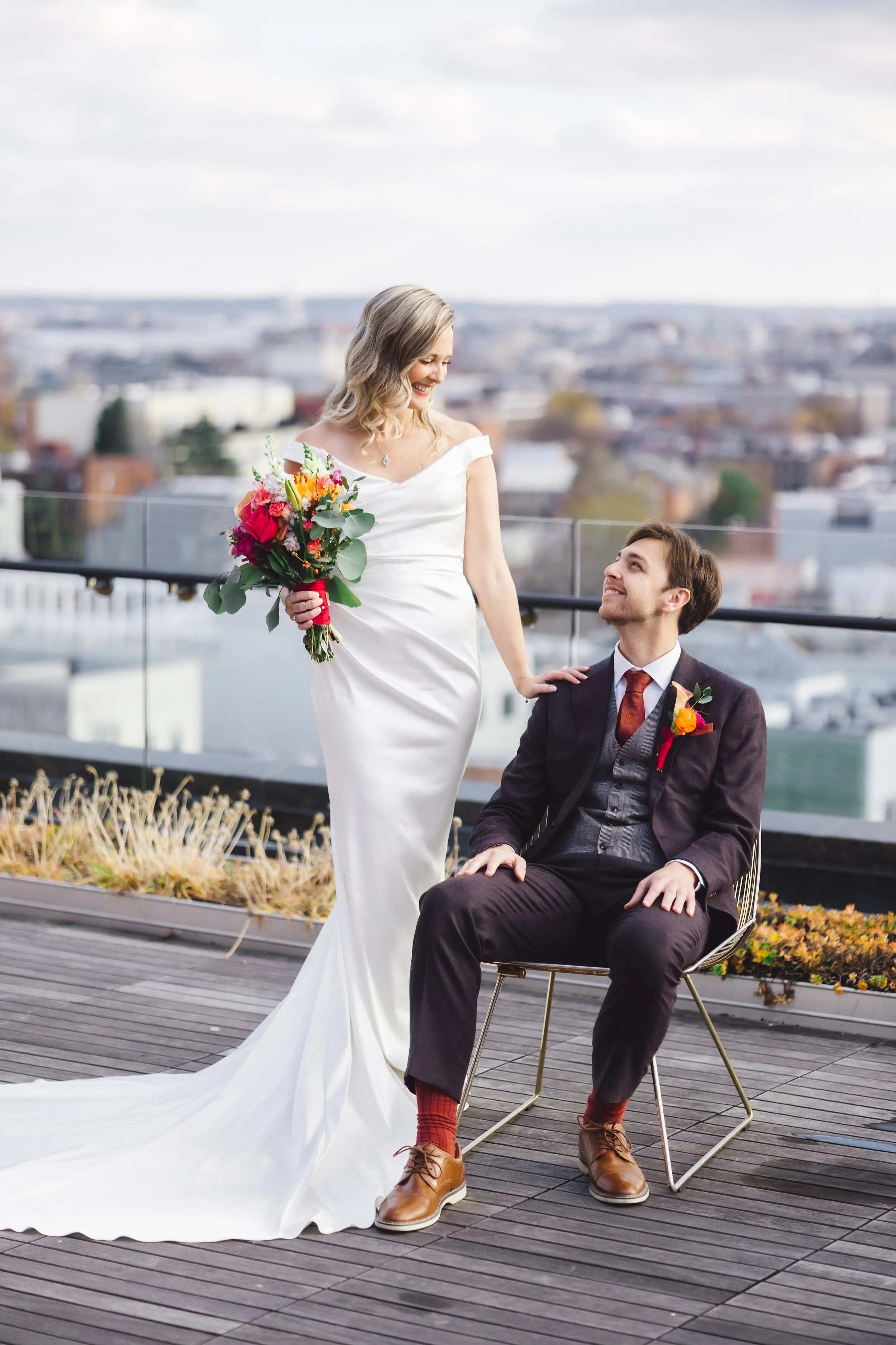 A bride in a white wedding gown standing next to a groom in a suit, seated on a chair, holding a bouquet of flowers, on a rooftop with cityscape in background.