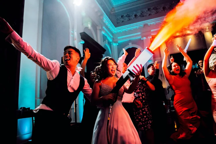 People dancing and celebrating at a party with colorful lighting, a woman holding a smoke or fire effect device, and joyful expressions.