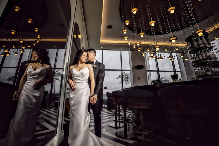 A bride and groom holding hands, standing next to a large mirror in a modern, stylish indoor space with large windows and unique lighting fixtures.
