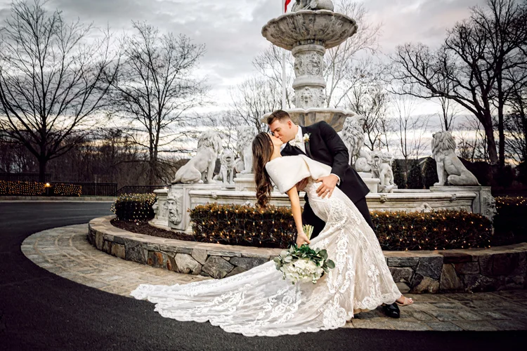 A newlywed couple shares a romantic kiss in front of a decorative fountain with lion statues at dusk. The bride is bending back in a white wedding gown, holding a bouquet, while the groom is dressed in a black tuxedo, holding her waist.