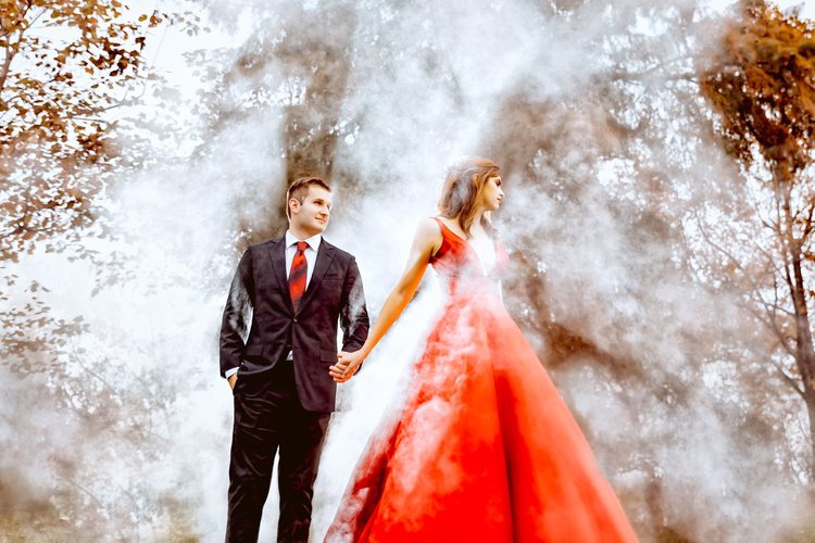 A couple in formal attire holding hands outdoors, with smoke or fog around them and trees in the background. The woman is in a red dress, and the man is in a suit.
