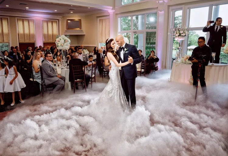 Wedding reception with bride and groom dancing in a cloud of smoke, guests seated at tables watching, and photographers capturing the moment.