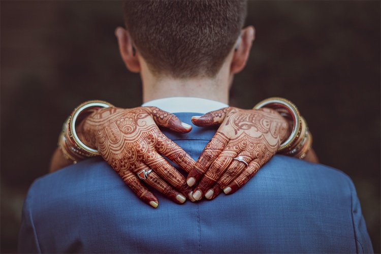 A person with henna-decorated hands hugging a man from behind, seen from behind, with a dark background.
