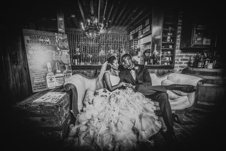 Black and white photo of a bride and groom sitting closely on a vintage sofa in a rustic bar, with the bride wearing a wedding dress and veil and the groom in a tuxedo. The bride is holding a bouquet, and they are gazing into each other's eyes. There