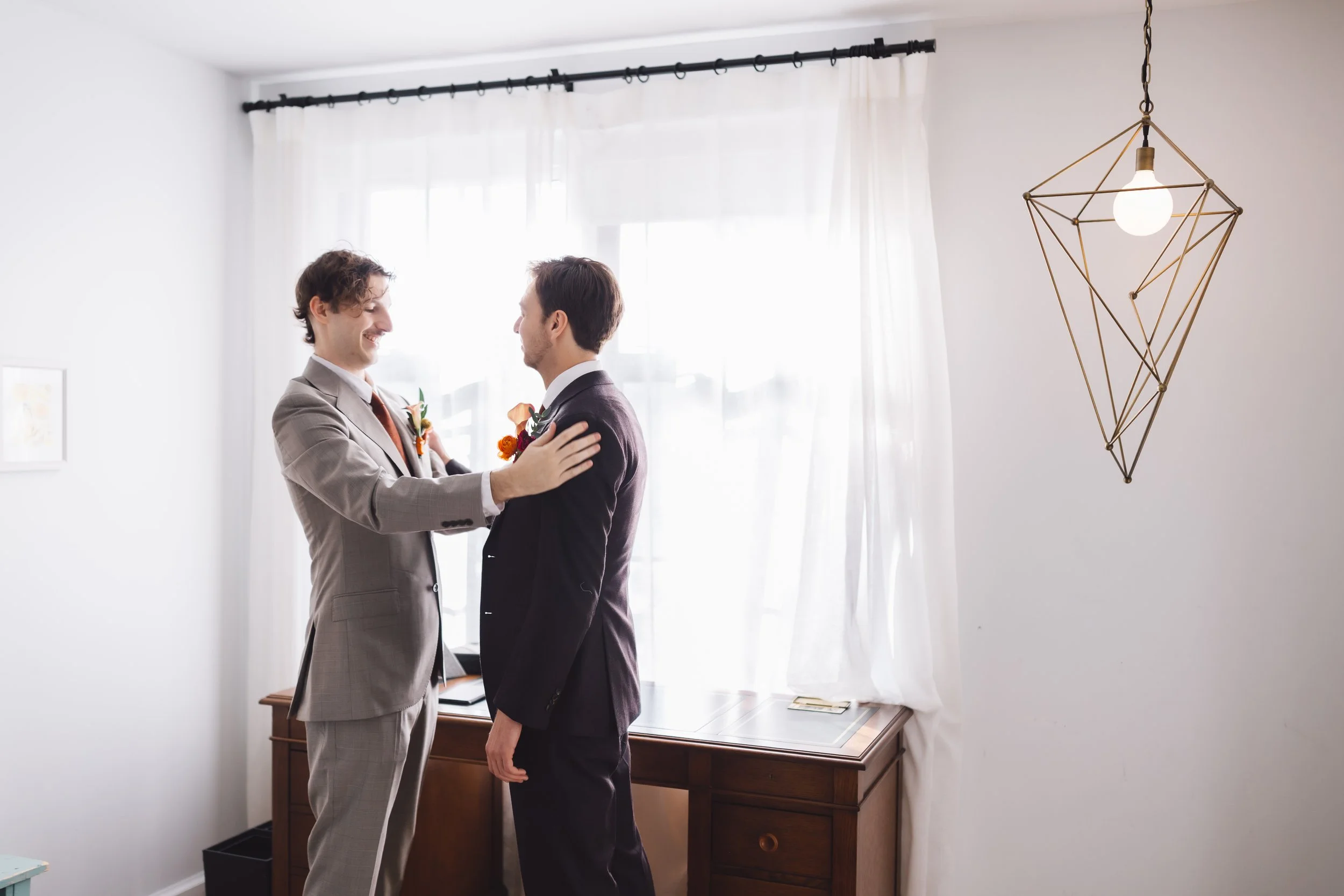 Two men in suits smiling and embracing in a bright room with white curtains and minimalistic decor.