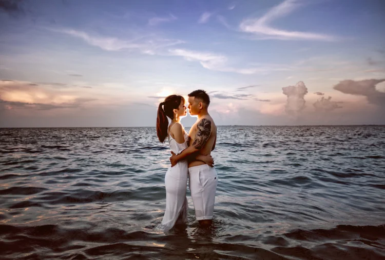 A couple standing in the ocean at sunset, facing each other close, with the sky and clouds in the background.