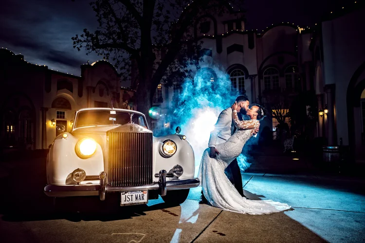A married couple on their wedding night sharing a kiss in front of a vintage white car, with a blue smoke effect behind them, in a venue with large windows and decorative lighting.