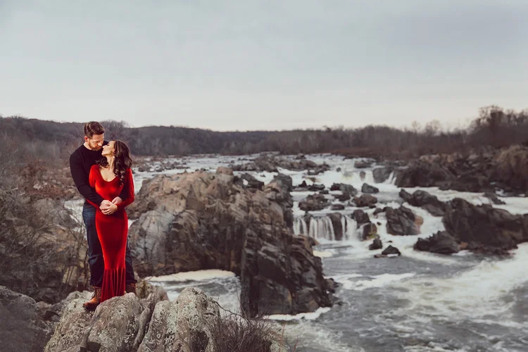 A couple standing on rocks by a river with waterfalls, the man wearing a black sweater and the woman wearing a long red dress, embracing and looking at each other.