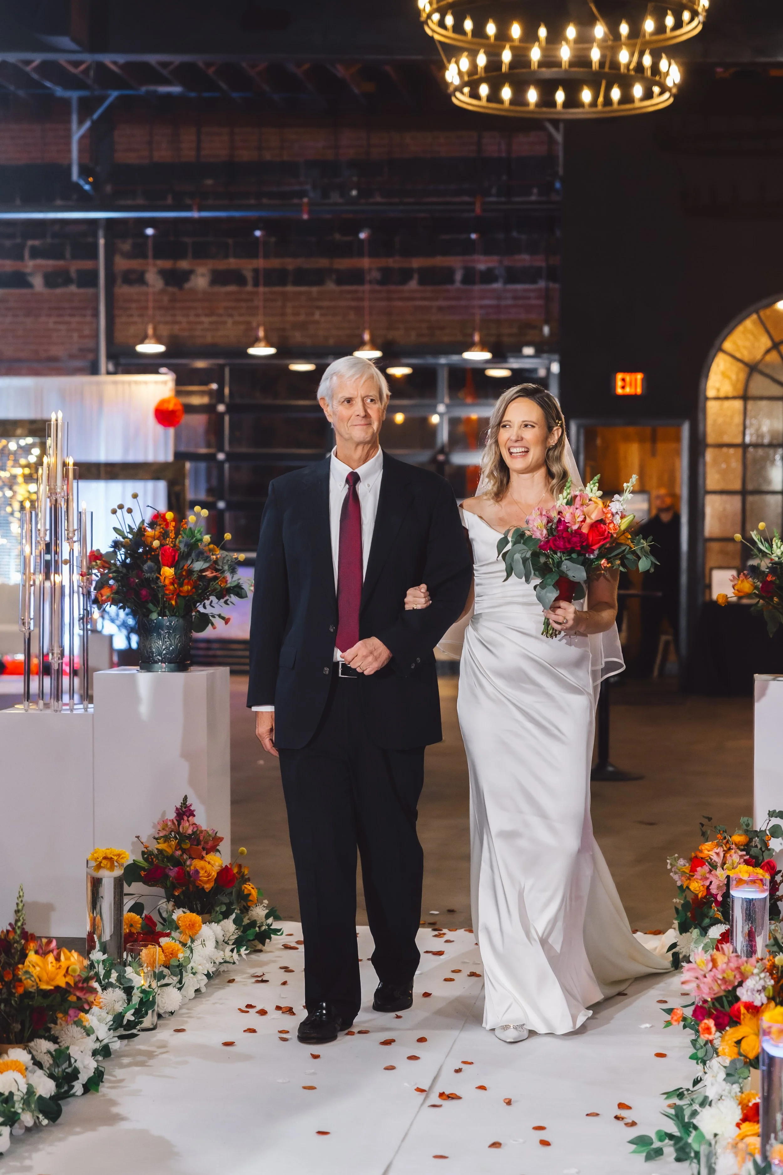 A bride in a white wedding dress holding a bouquet walking down the aisle with her father in a dark suit and red tie, in a decorated wedding venue with floral arrangements and candles.