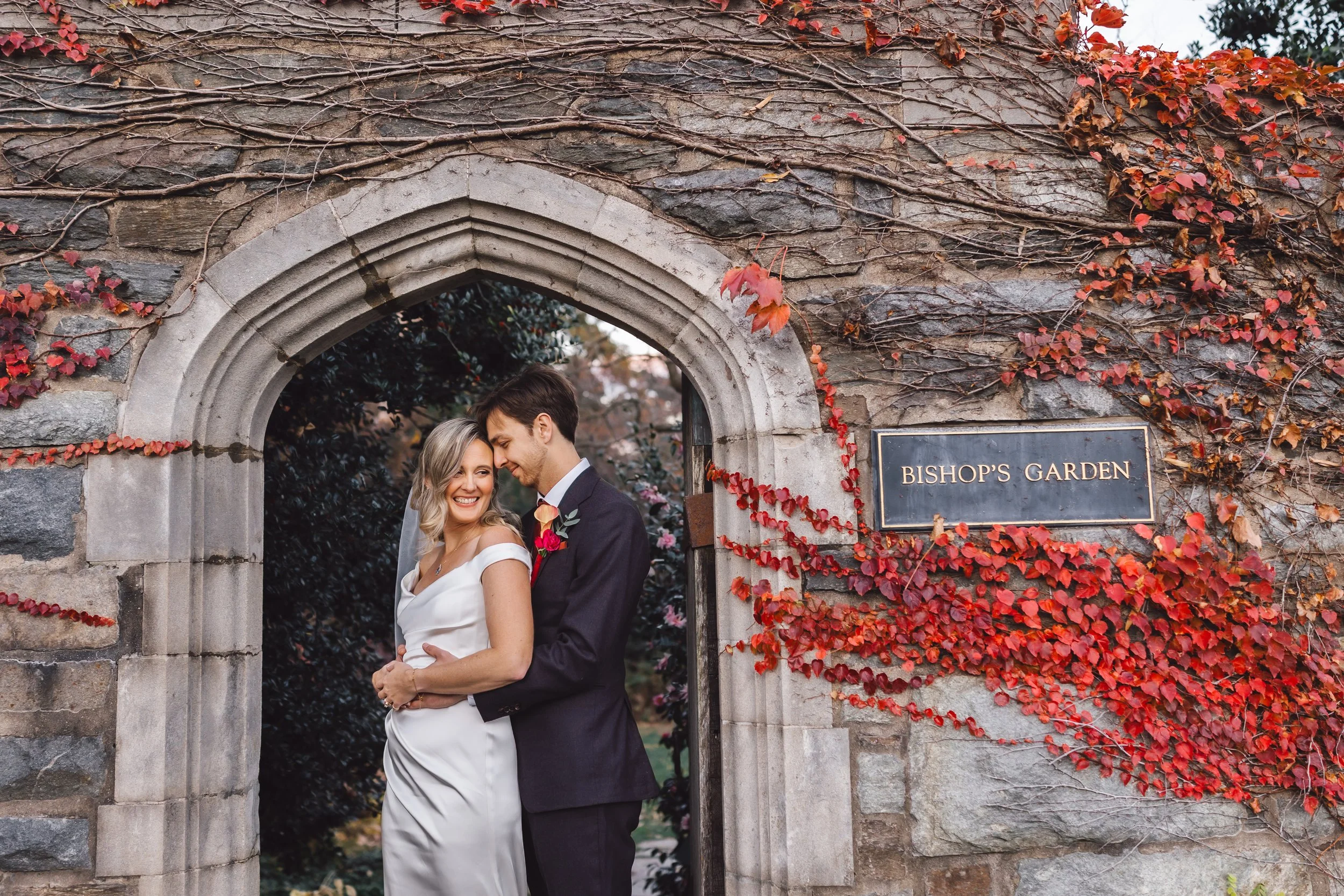A newlywed couple standing in an arched stone doorway decorated with red and orange autumn leaves, smiling and holding each other, with a sign that reads 'Bishop's Garden' on the wall to the right.