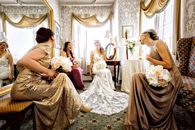 A bride in a white wedding dress sitting and laughing in a vintage room with five women around her, all holding bouquets and dressed in elegant gowns, sharing a joyful moment.