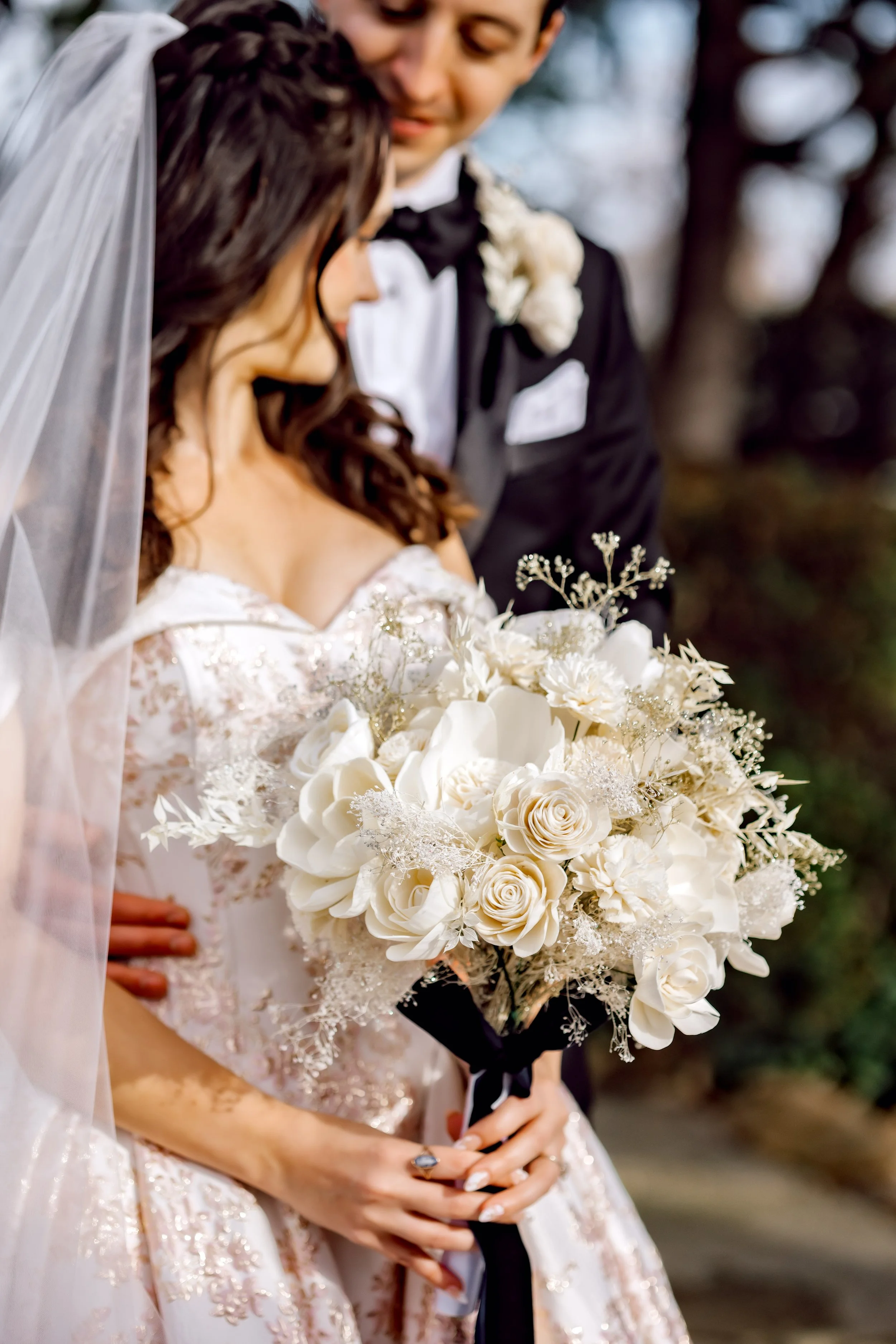 A bride and groom standing close together outdoors, with the bride holding a large bouquet of white flowers.