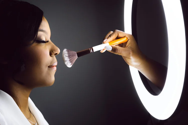 A woman with makeup on her face, including eyeliner, eyeshadow, and lipstick, is having makeup applied with a large makeup brush, held by a person with glittery nail polish, in front of a mirror with round light.