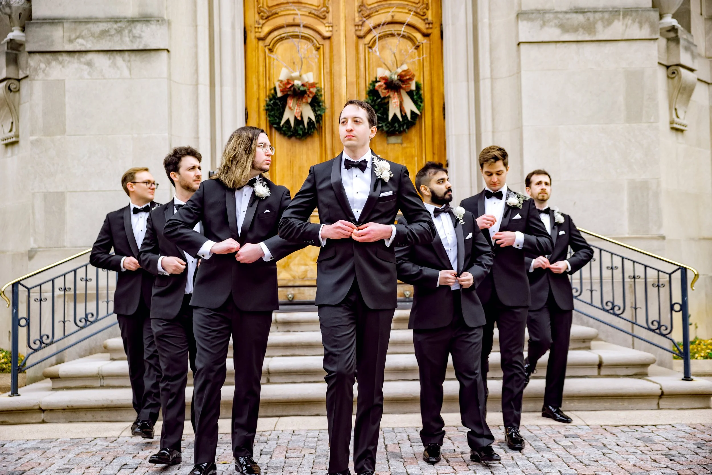 Group of men in tuxedos standing on steps outside a building with decorated doors.