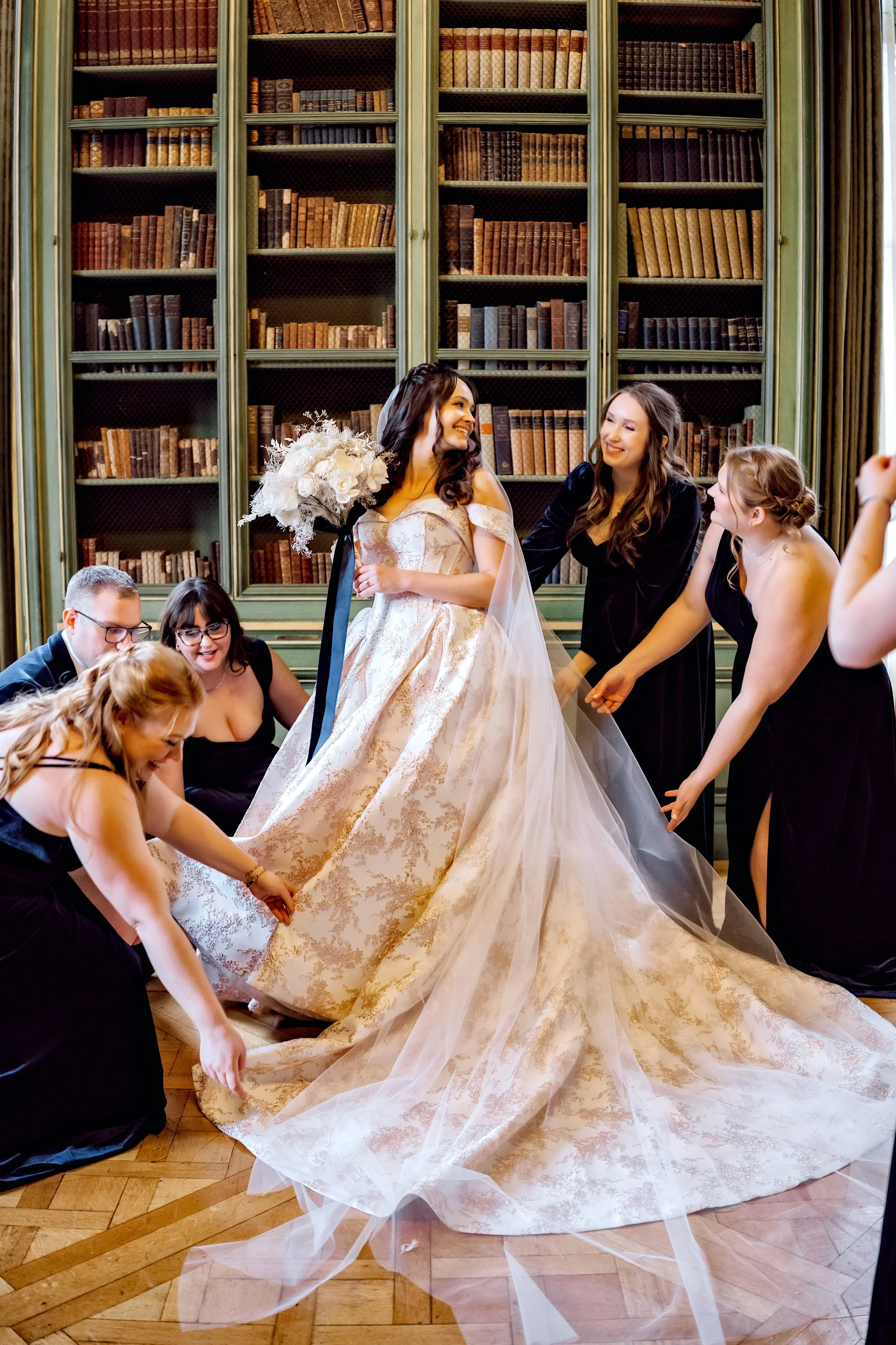 Bride in a gold and white wedding gown surrounded by bridesmaids and friends in a library room with green bookshelves.