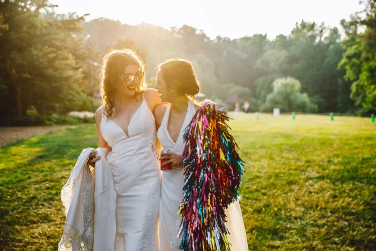 Two women in white wedding dresses sharing a joyful moment outdoors at sunset. One woman holds a colorful tinsel decoration, and both are smiling and appear to be celebrating.