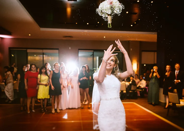 Bride in a white lace gown throwing her bouquet into the air at her wedding reception, surrounded by guests in a decorated indoor venue.