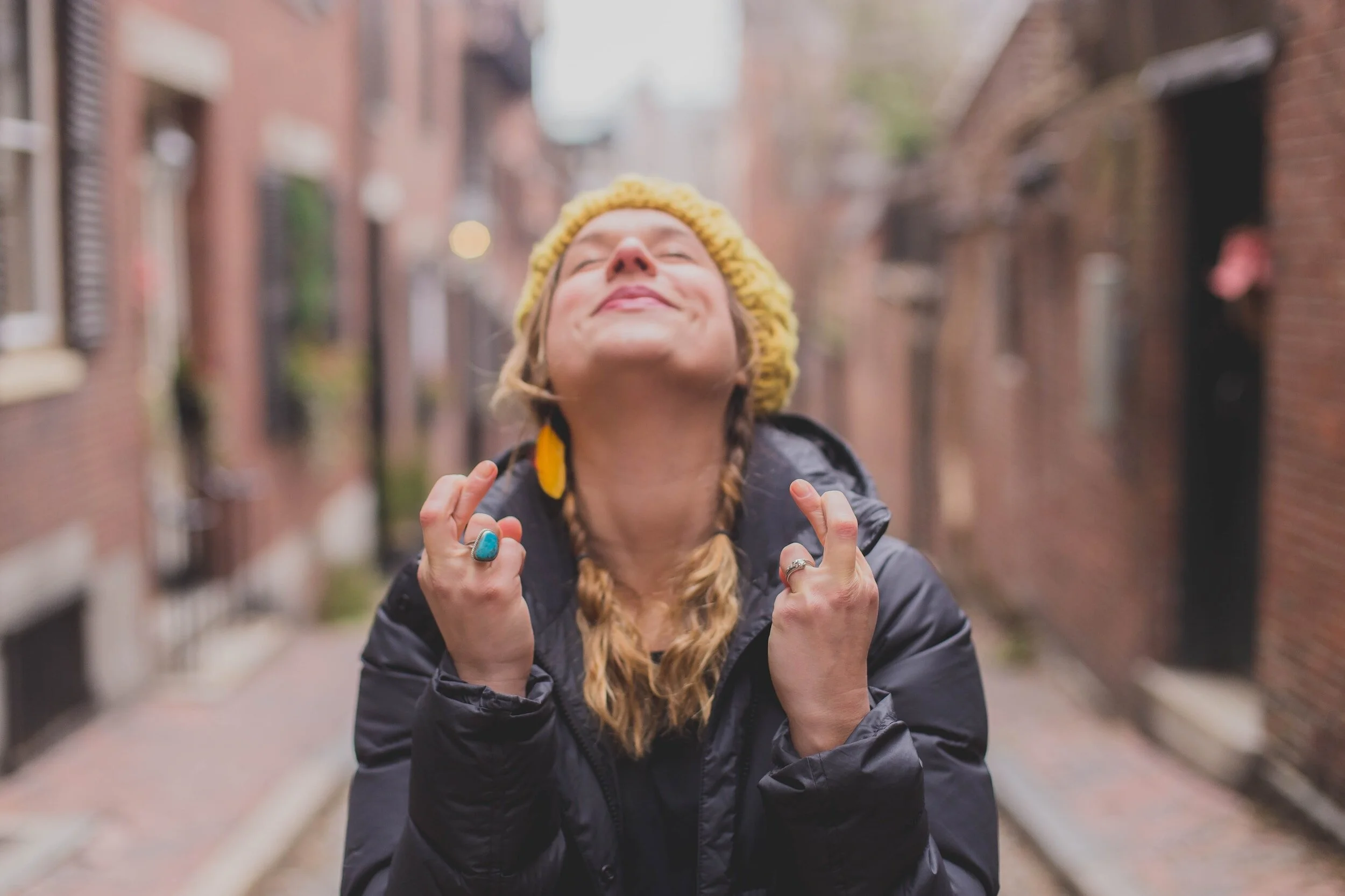 Woman with braided hair, wearing a yellow beanie and a black jacket, standing on a brick alleyway, smiling with her eyes closed and fists clenched in excitement.