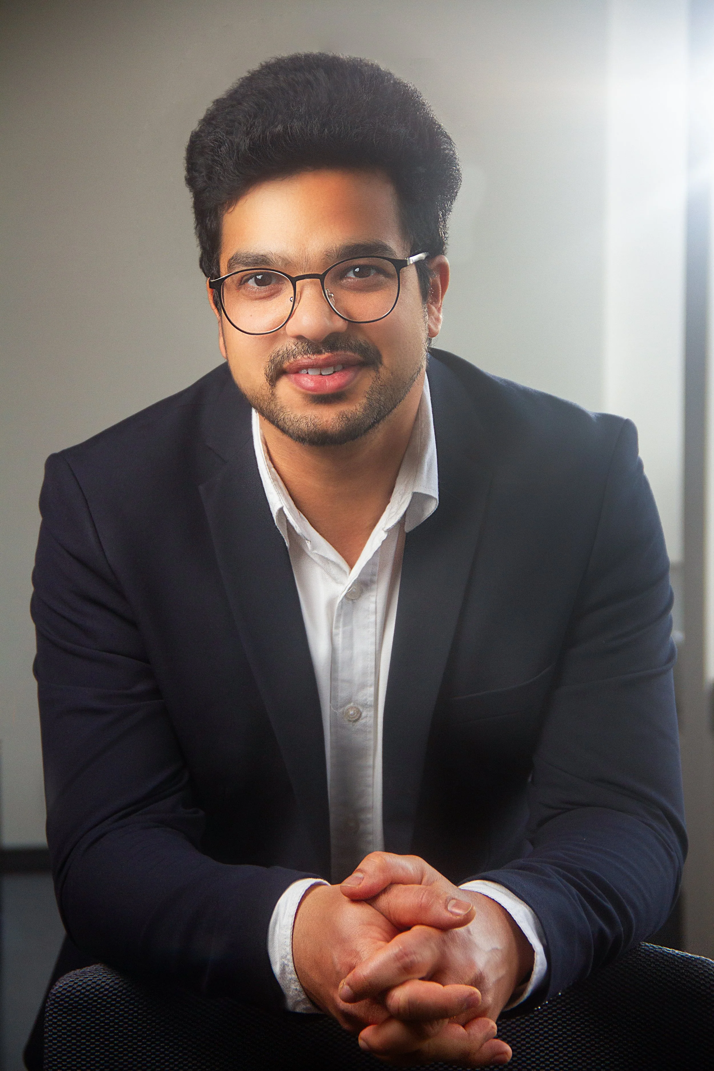 A man with dark hair, glasses, and a beard, dressed in a suit and white shirt, sitting with hands clasped together in a professional setting.