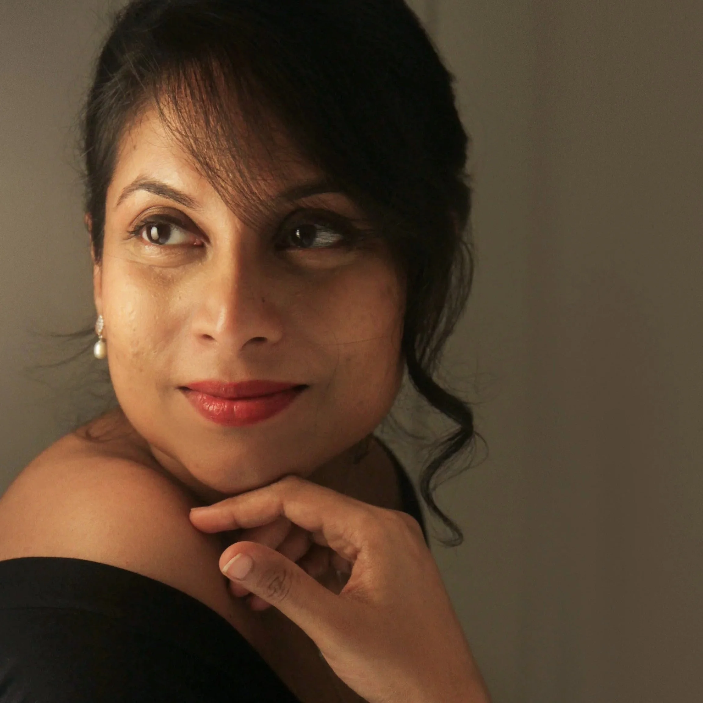 A woman with short dark hair, wearing earrings and makeup, smiles subtly while resting her chin on her hand against a plain background.