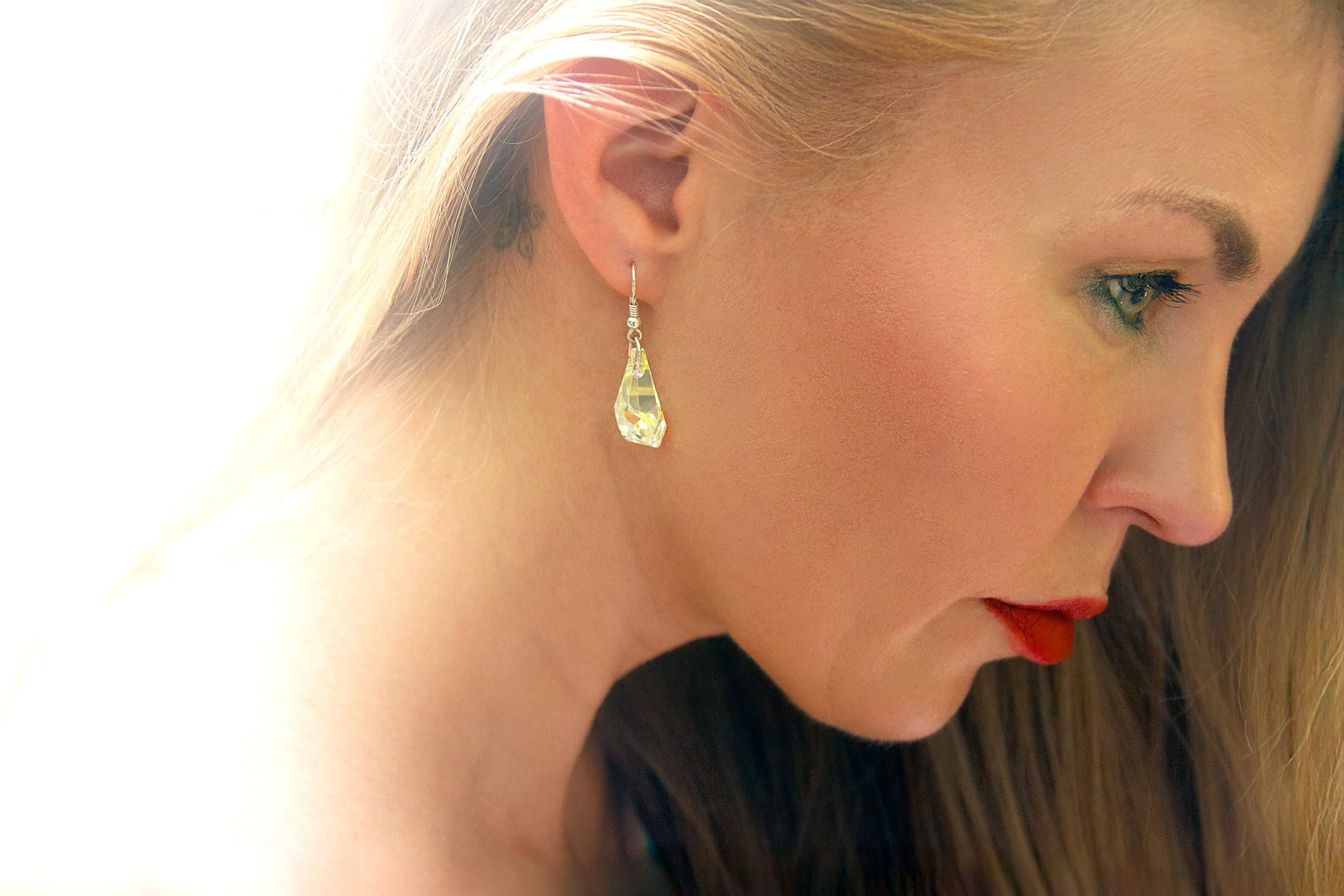 Close-up side profile of a woman with blonde hair, wearing a sparkling drop earring and red lipstick, looking downward with a soft expression.