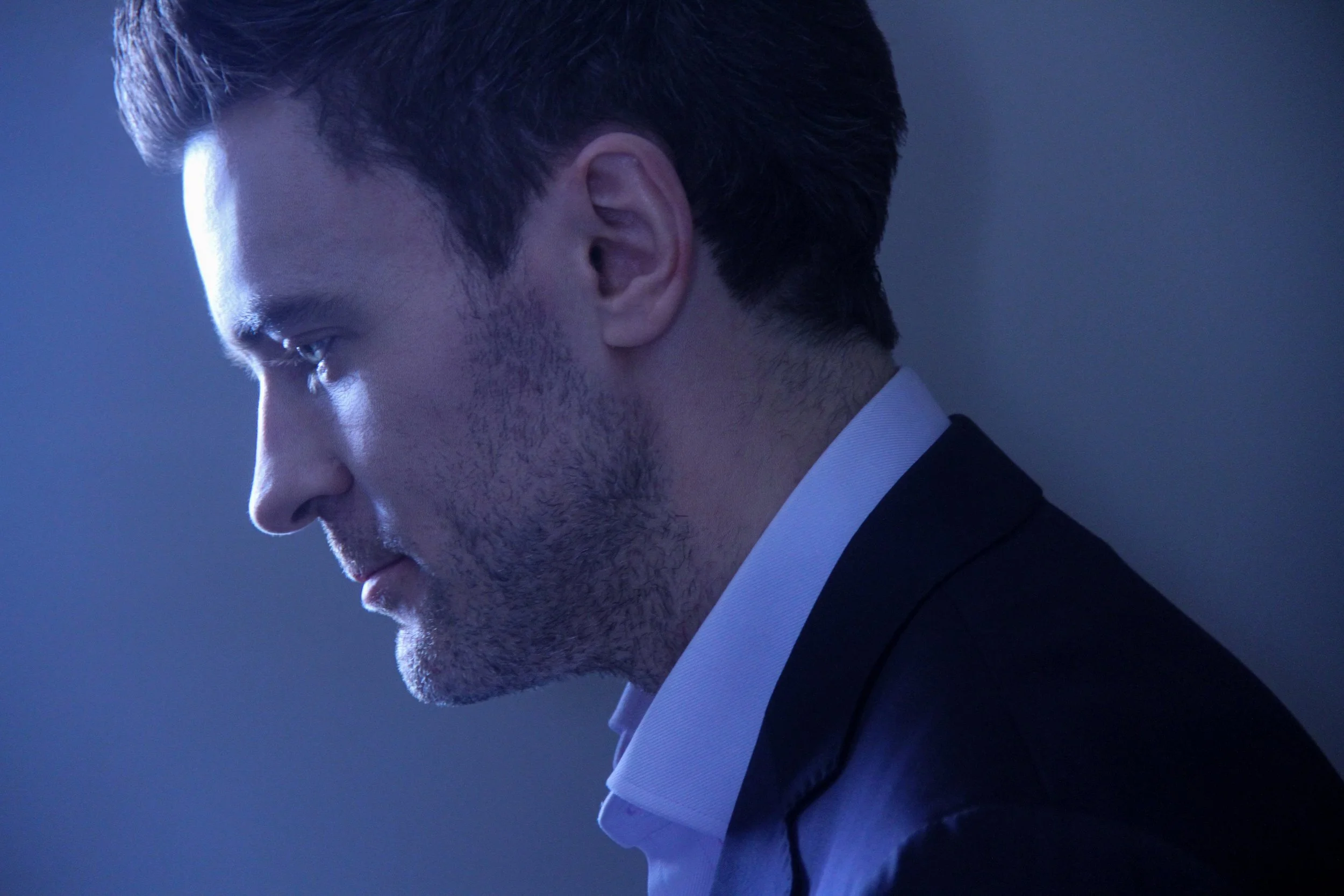 Close-up side profile of a man with dark hair and stubble, wearing a white shirt and black suit, looking downward against a gray background.