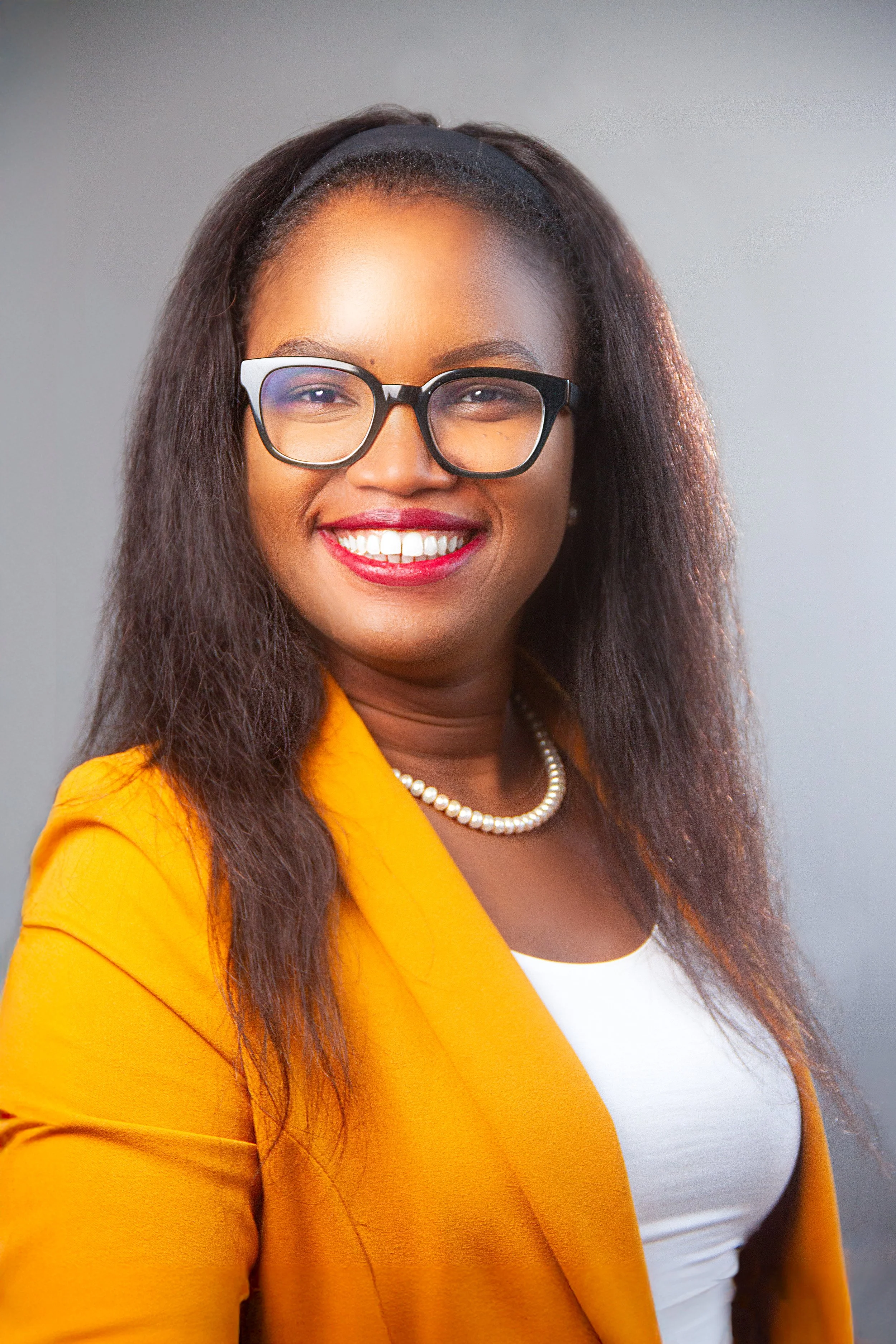 A smiling woman with glasses wearing a yellow blazer and white top, with a pearl necklace, against a neutral background.