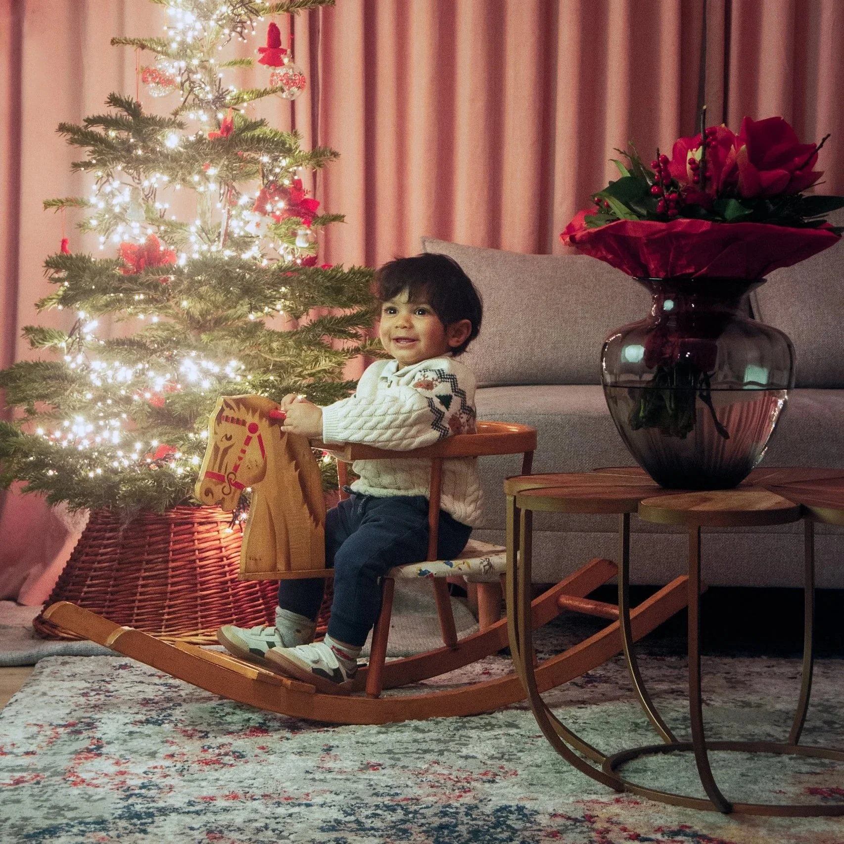 A young boy sitting on a wooden rocking horse in a living room decorated for Christmas, with a lit Christmas tree and a vase of red flowers on a wooden table nearby.