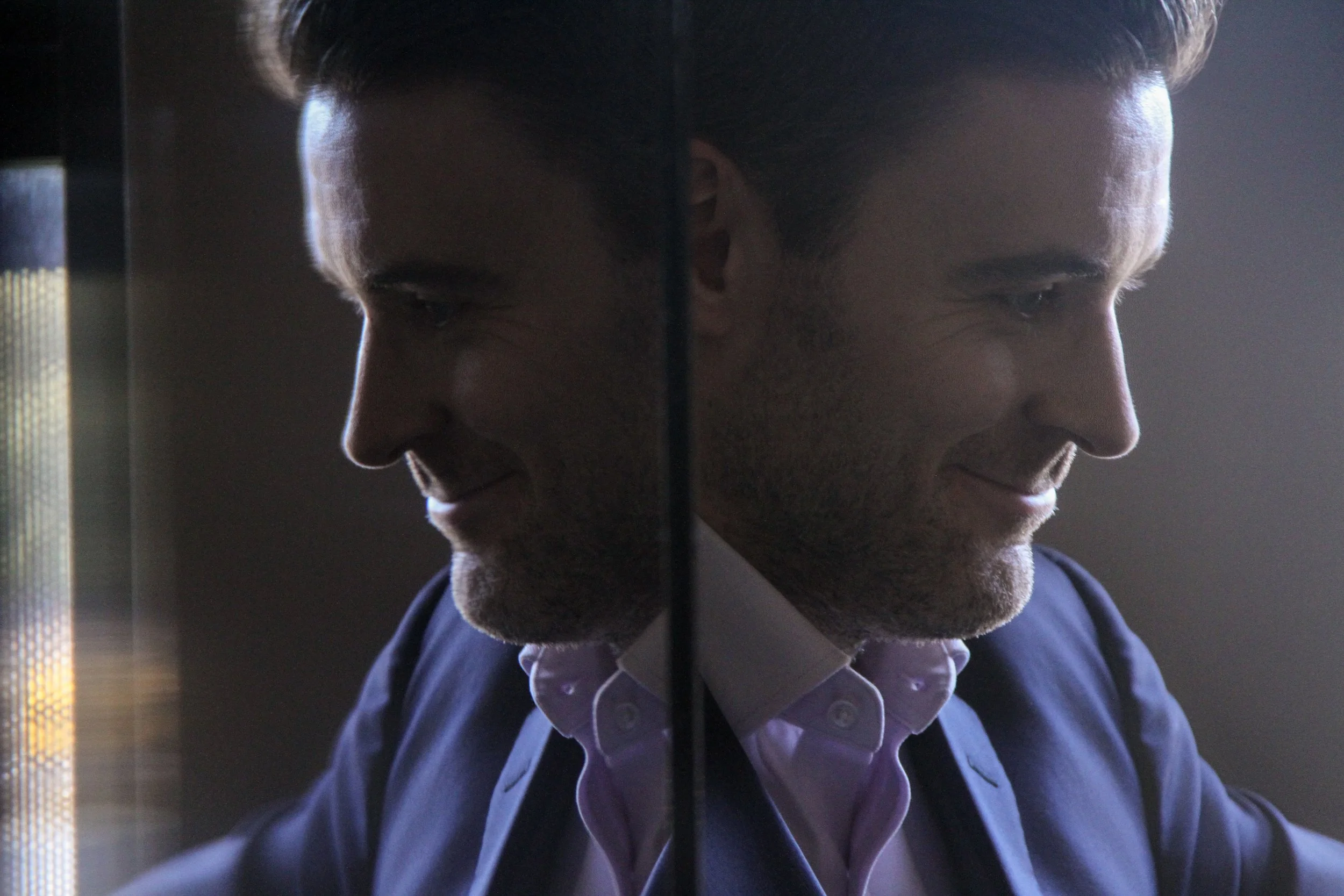 Side profile of a man with slicked back hair, wearing a suit and white shirt, smiling slightly, with his reflection visible on a mirror or glass surface.