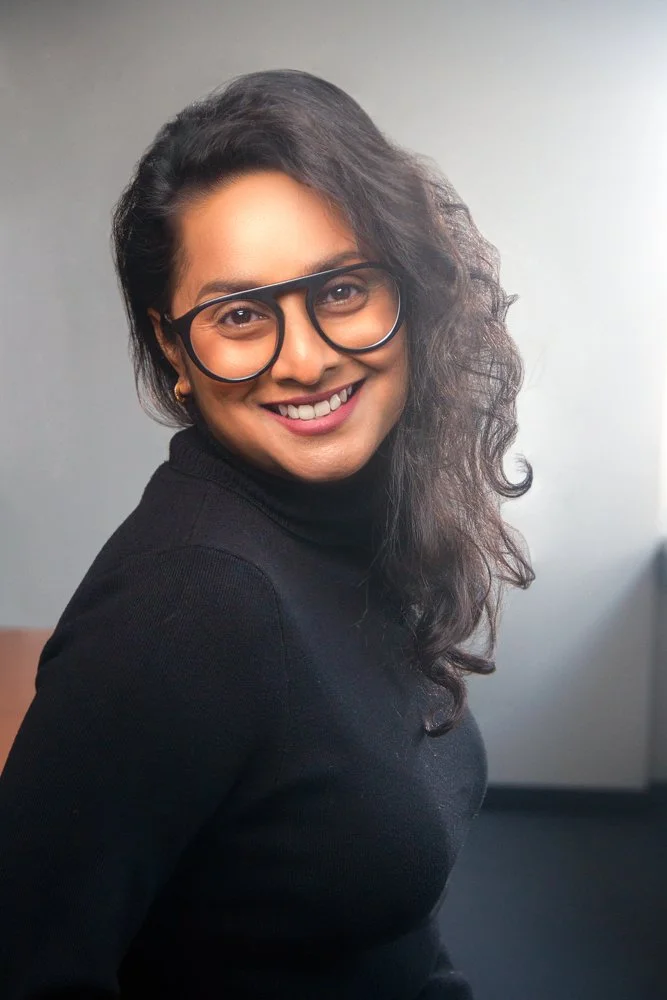 A woman with curly hair wearing glasses and a black turtleneck, smiling at the camera against a neutral gray background.