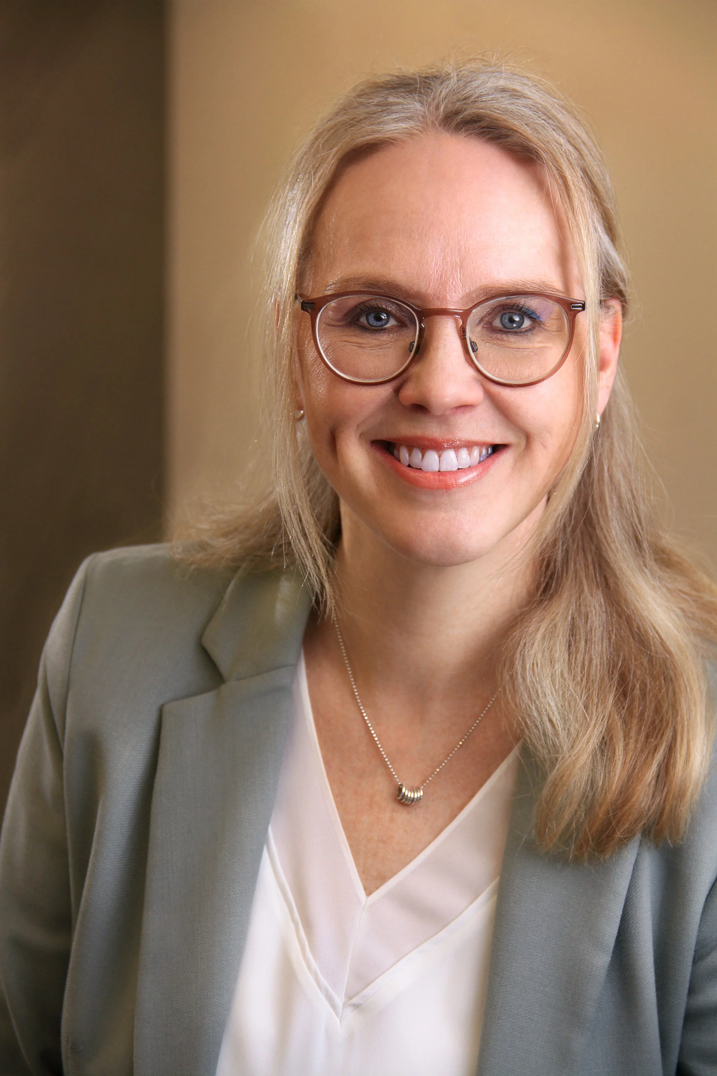 A woman with blonde hair wearing glasses, a light gray blazer, and a white blouse, smiling at the camera.
