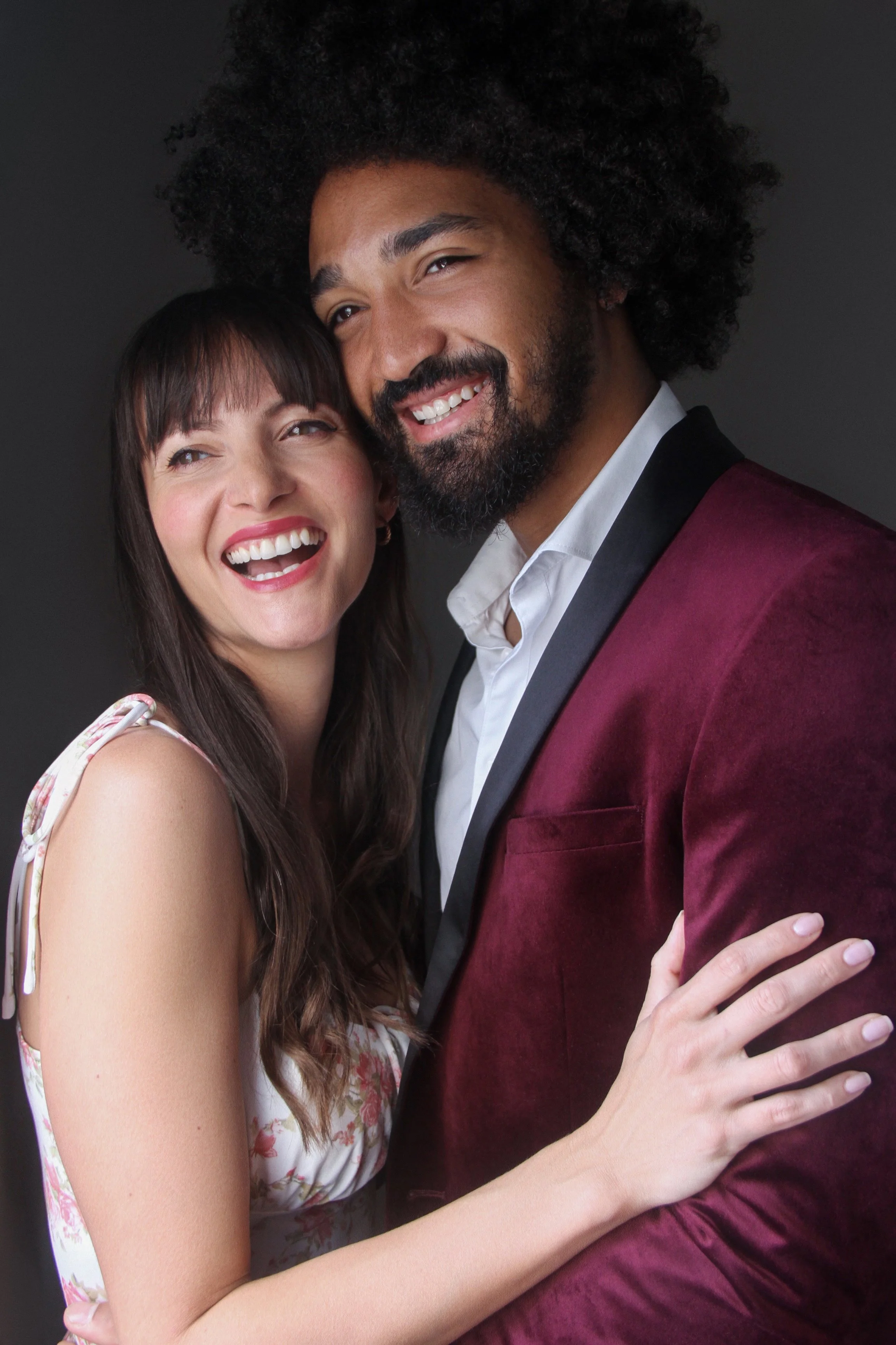 A happy couple embracing, smiling, in front of a dark background. The woman has long brown hair and wears a sleeveless floral dress. The man has curly hair and a beard, and wears a maroon velvet blazer with black lapels and a white shirt.
