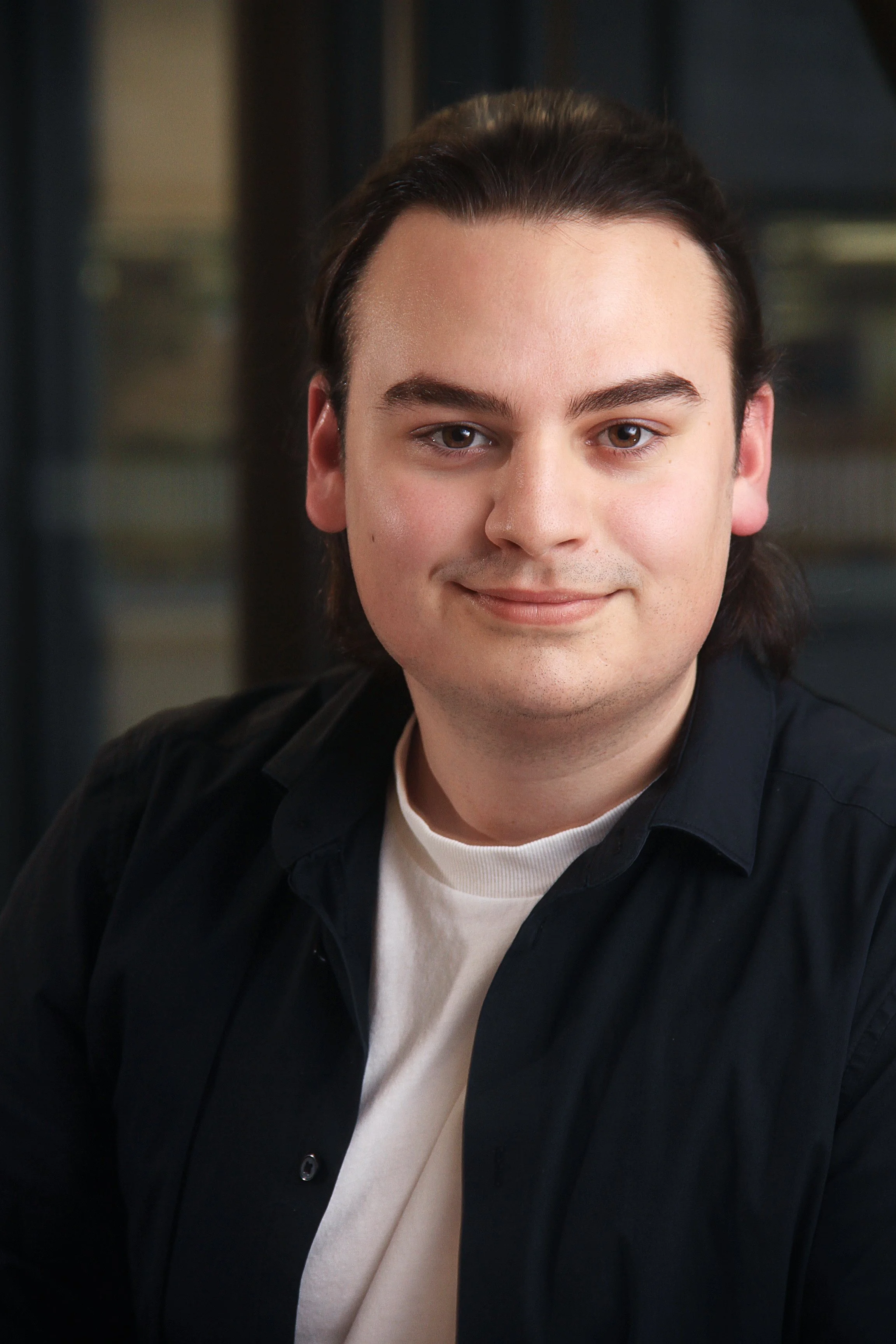 A young man with fair skin, dark brown hair, and brown eyes, wearing a black shirt over a white t-shirt, smiling softly indoors.