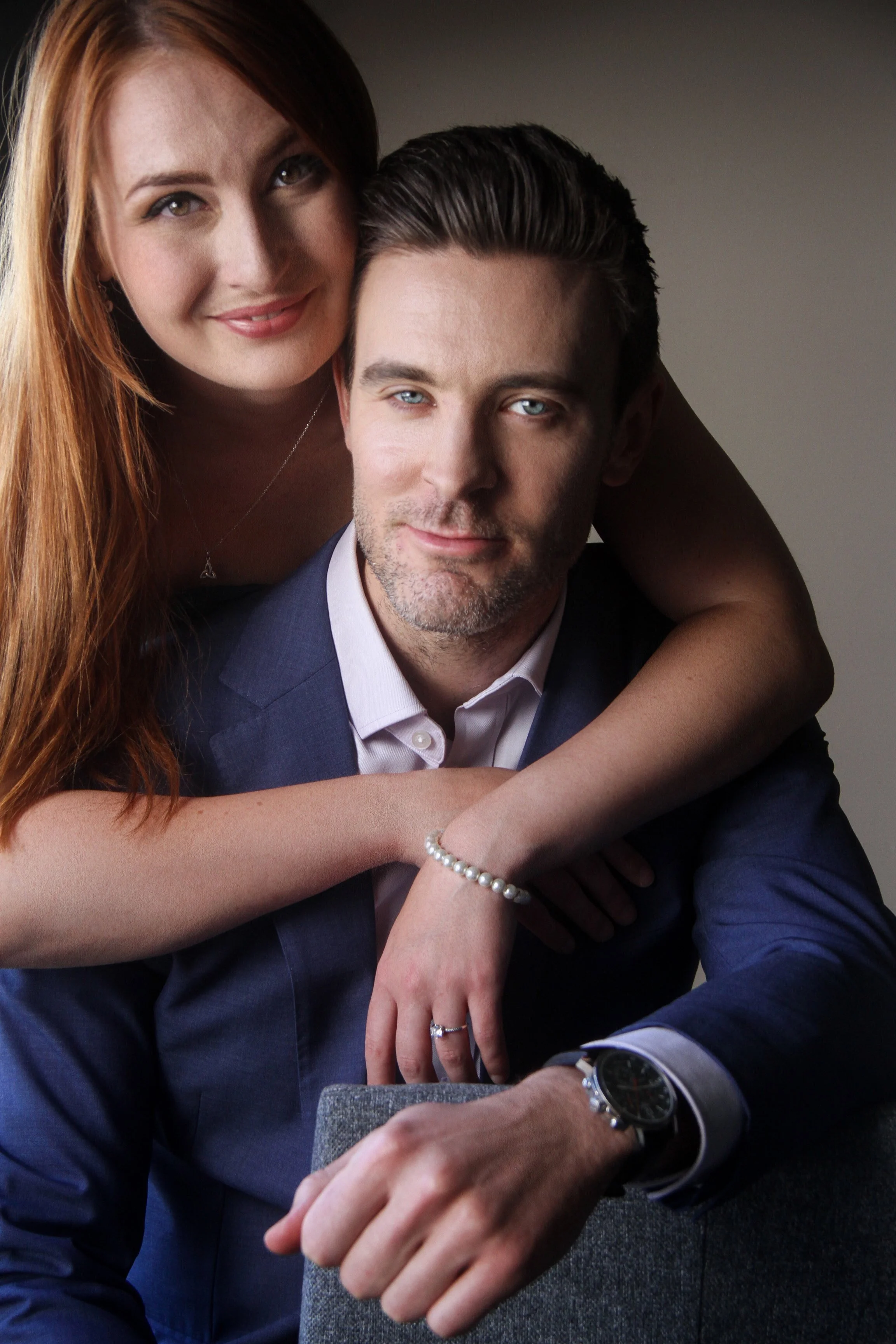 A woman with long red hair leaning on a man with dark hair, both wearing formal attire, with the woman smiling and the man looking at the camera.