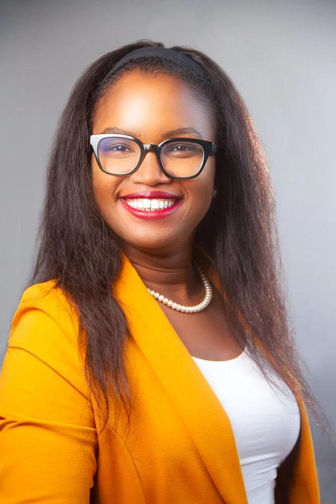A woman with long dark hair, glasses, and a bright smile, wearing a yellow blazer, a white top, and a pearl necklace, poses against a gray background.