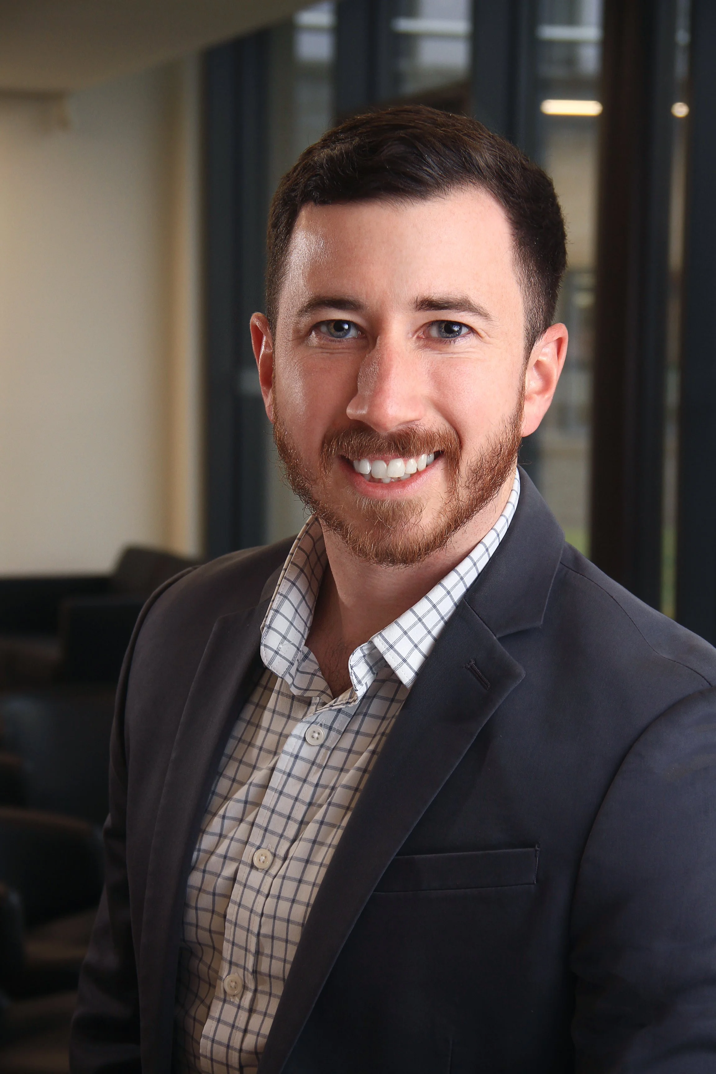 A man with brown hair, a beard, and blue eyes smiling wearing a dark blazer and a checked shirt in an office setting.