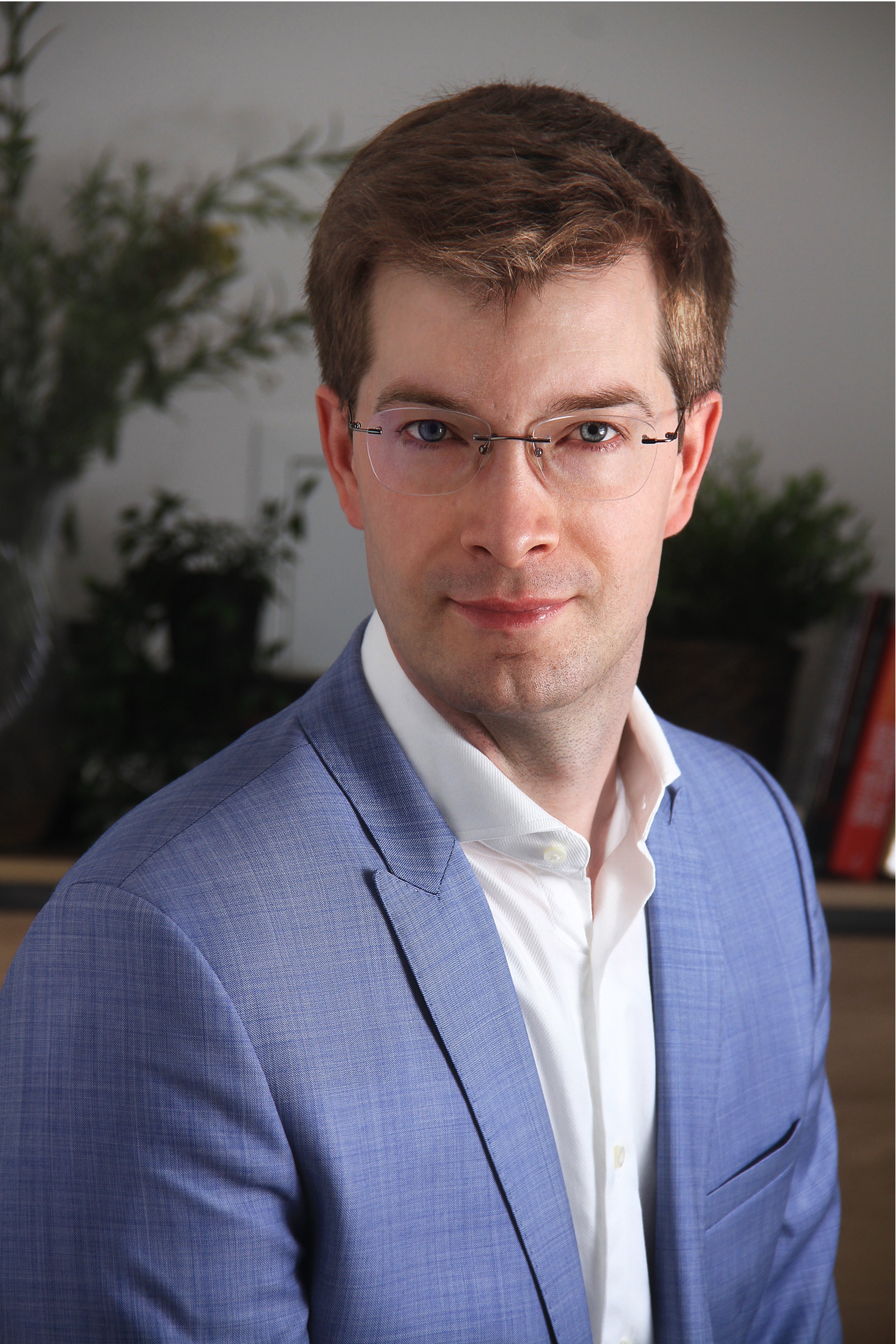 A man with short brown hair wearing glasses, a white shirt, and a blue blazer in an office setting with plants in the background.