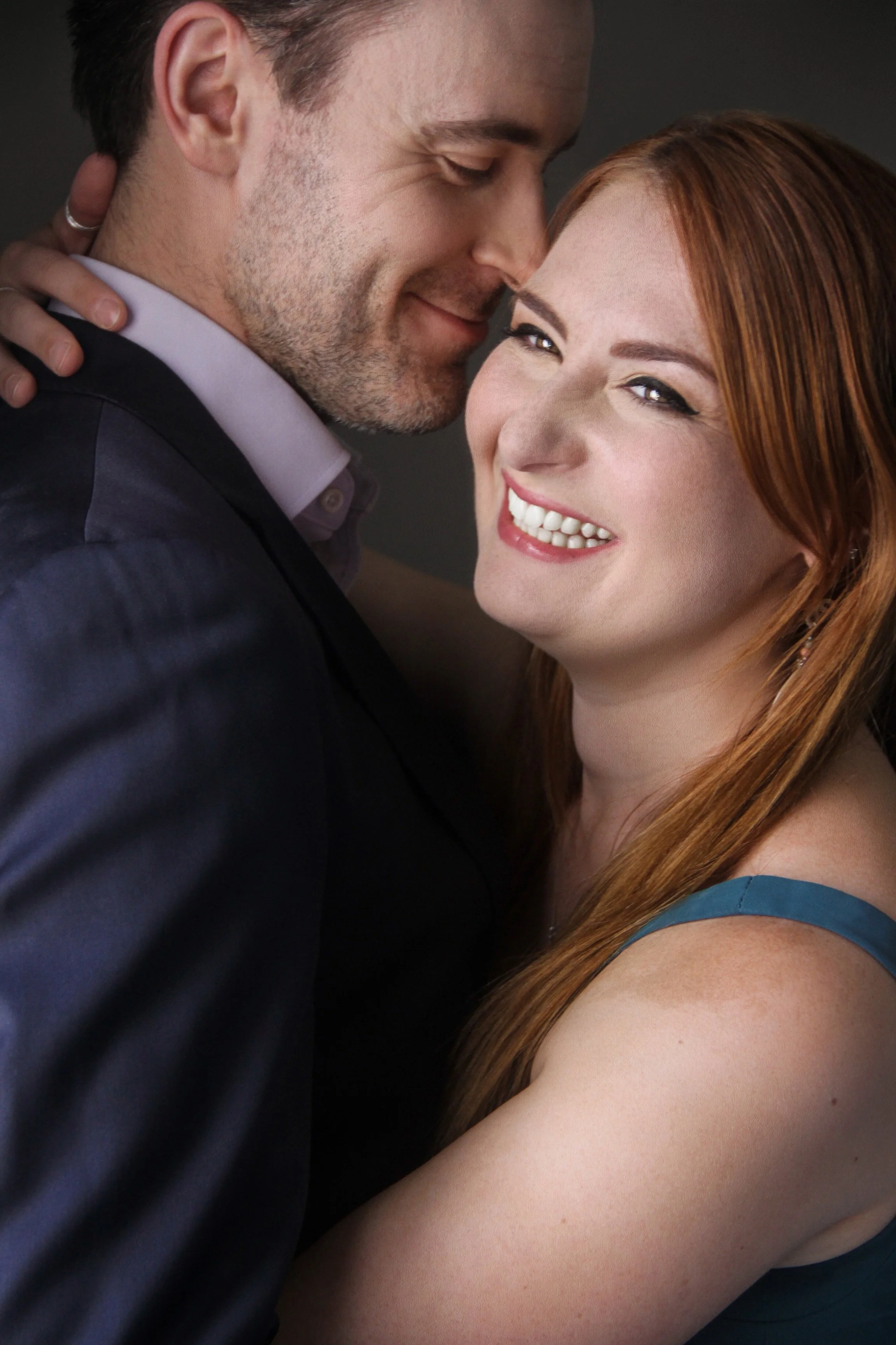 A smiling red-haired woman hugging a man with a slight beard, both dressed in formal attire, in a close embrace.