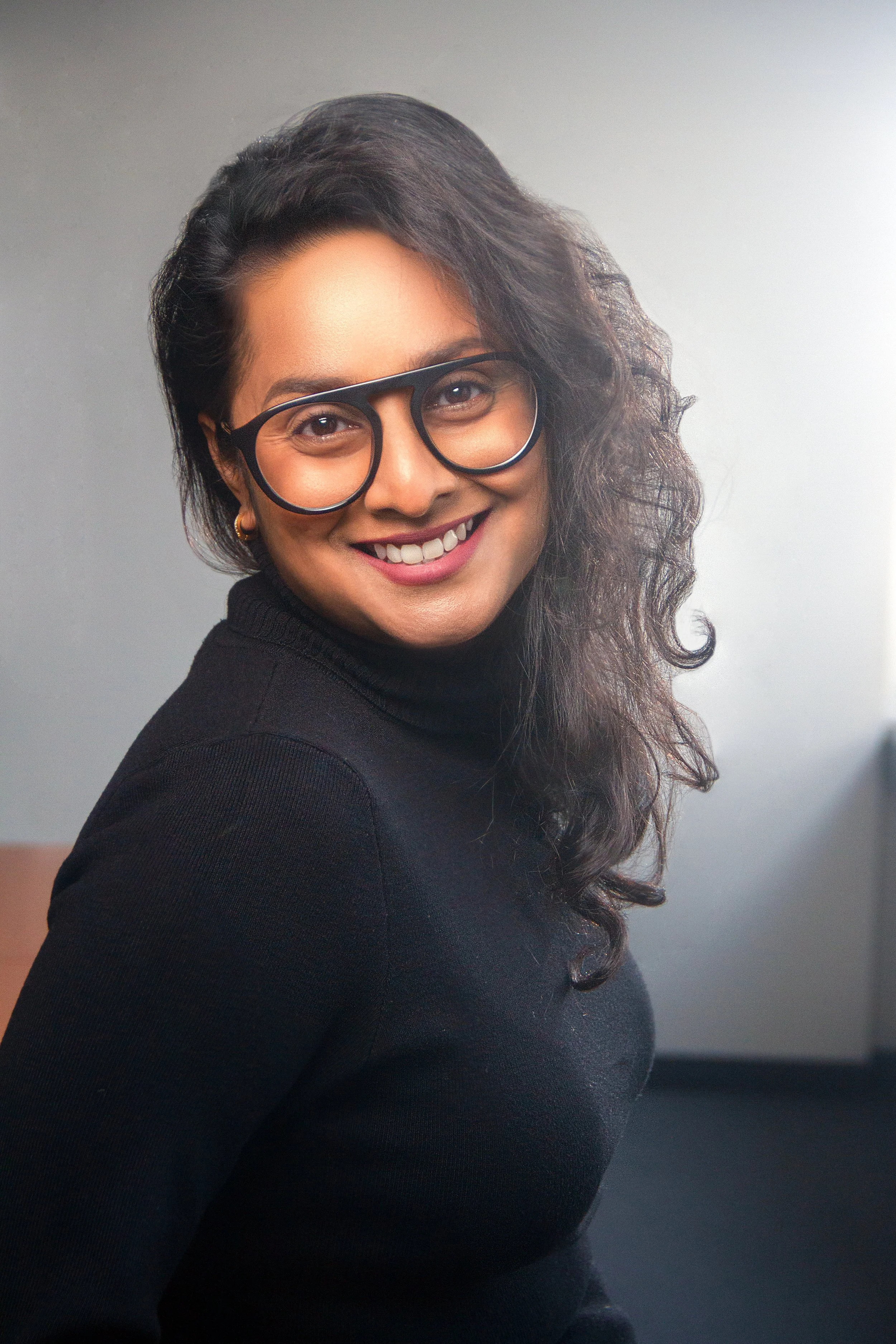 A woman with dark curly hair, wearing glasses and a black turtleneck sweater, smiling at the camera against a plain background.