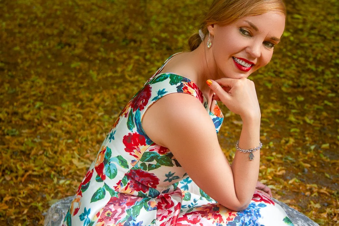 A woman with blonde hair styled in a low bun, wearing a colorful floral dress and silver jewelry, sitting on a rock in a forest with autumn leaves ground, smiling at the camera.