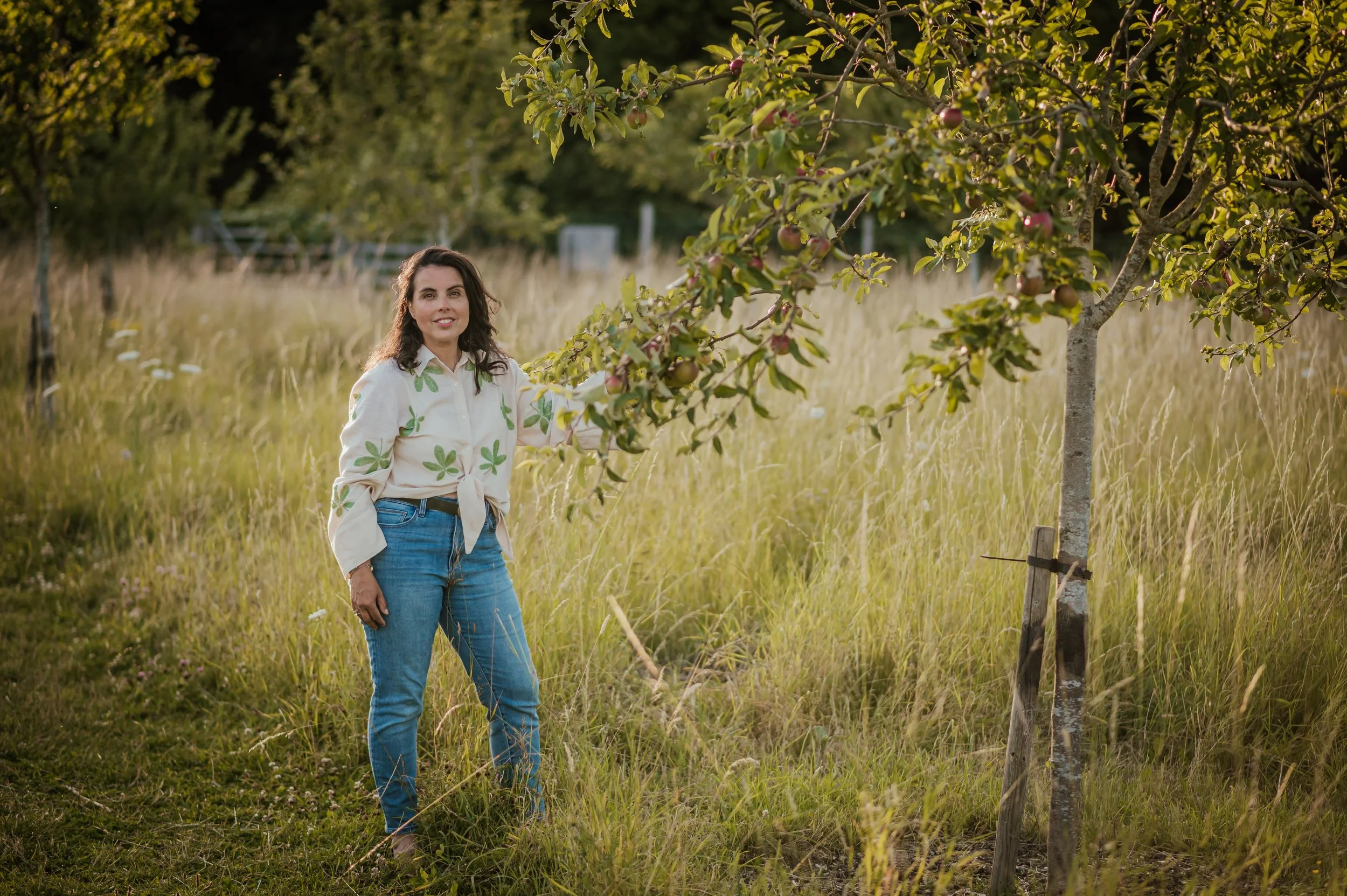 Garden designer in an apple orchard, touching a branch, wearing a light-colored top and jeans, surrounded by tall grass.