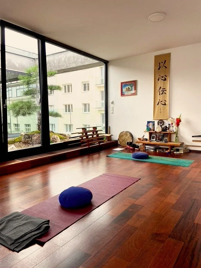 A yoga or meditation room with wooden floors, large windows overlooking an outdoor Garden, yoga mats with pillows, and spiritual or decorative items on a small shelf.