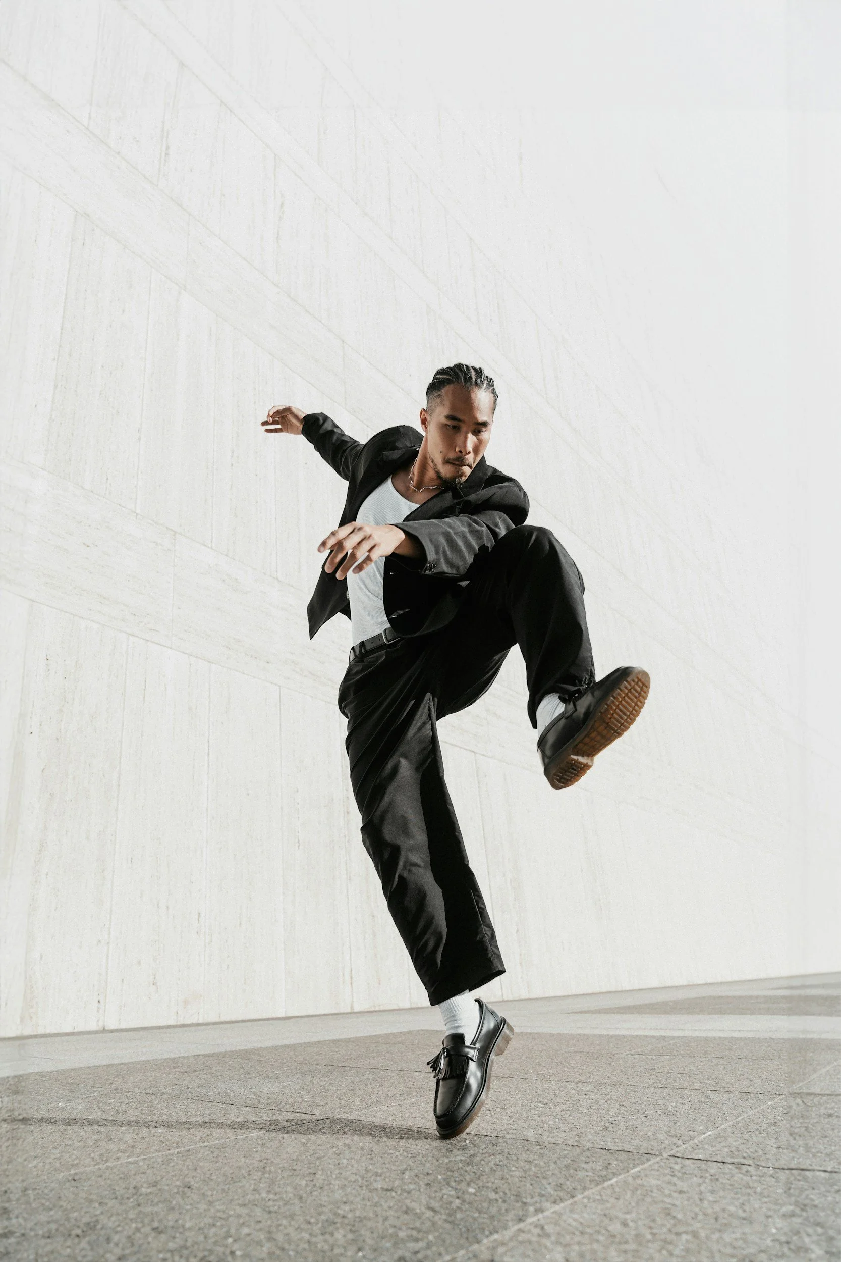 A man dressed in a black blazer, white shirt, and black pants performs a dance move or a high kick outdoors against a plain white wall. He has dark hair styled back and is wearing black shoes and white socks.