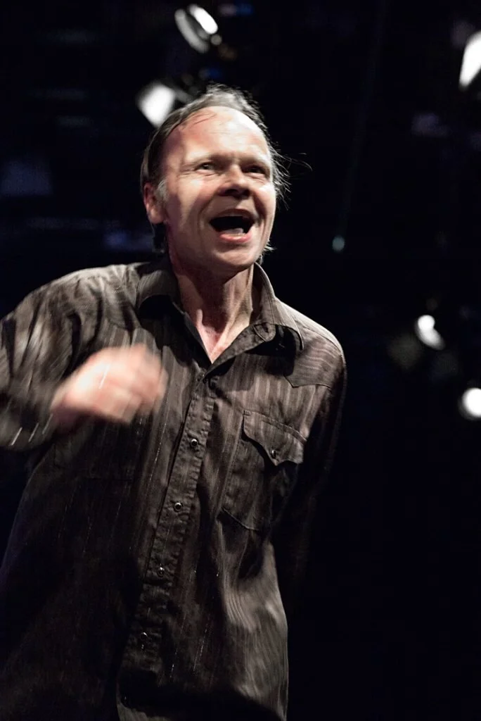 A man with light skin and short hair appears to be speaking or singing, with a dark background and stage lighting overhead.