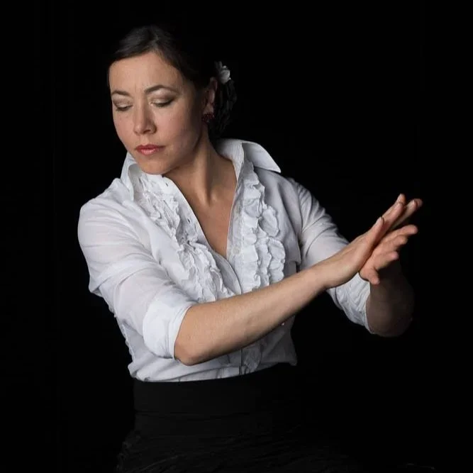 A woman with dark hair styled back, wearing a white blouse with ruffles, appears to be gesturing with her hands against a black background.