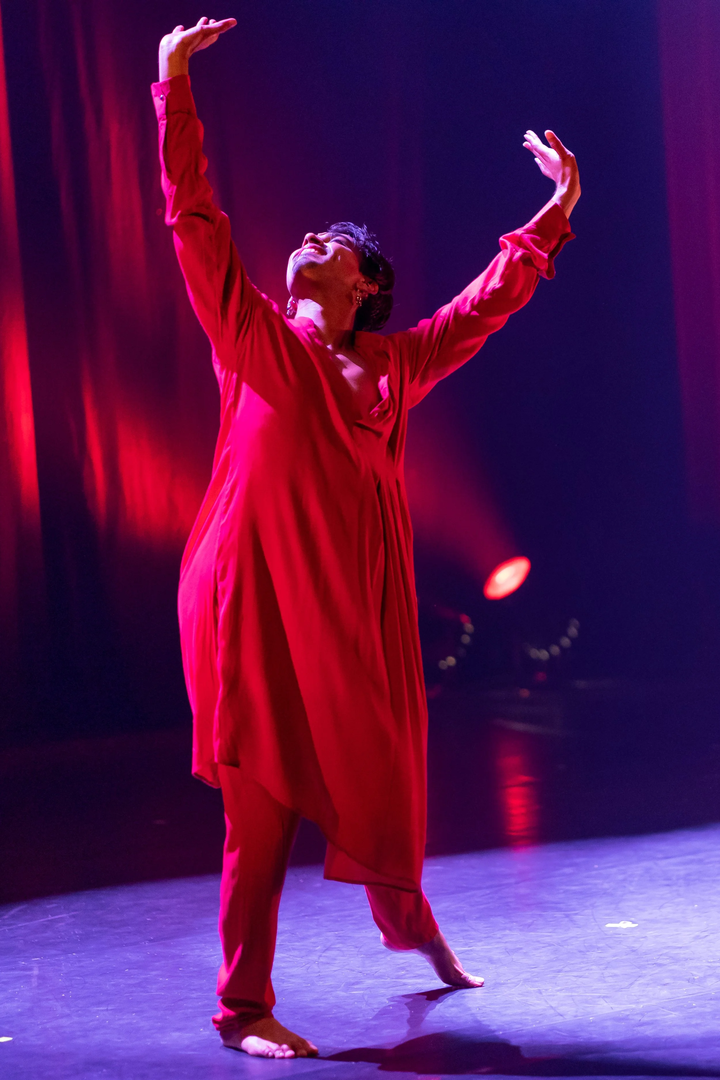 A woman in a red dress performing a dance with arms raised and head tilted back on a stage illuminated by red and purple lights.