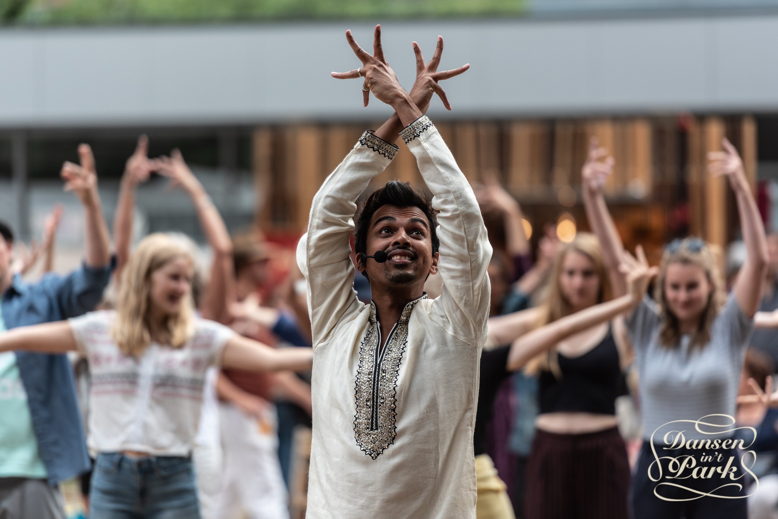 A dance instructor leads a group in an outdoor dance class. The instructor, wearing a white traditional outfit with embroidery, has their hands gracefully raised above their head. A diverse group of participants follows along in the background.