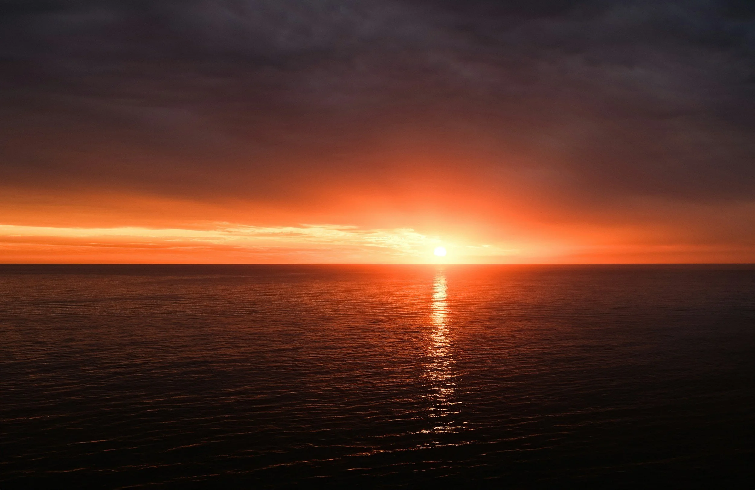 Sunset over the ocean with dark clouds in the sky and the sun partially visible near the horizon, reflecting on the water.
